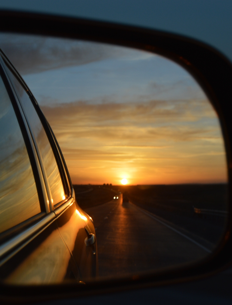 Car driving down an open road with the sunset in the rear view mirror
